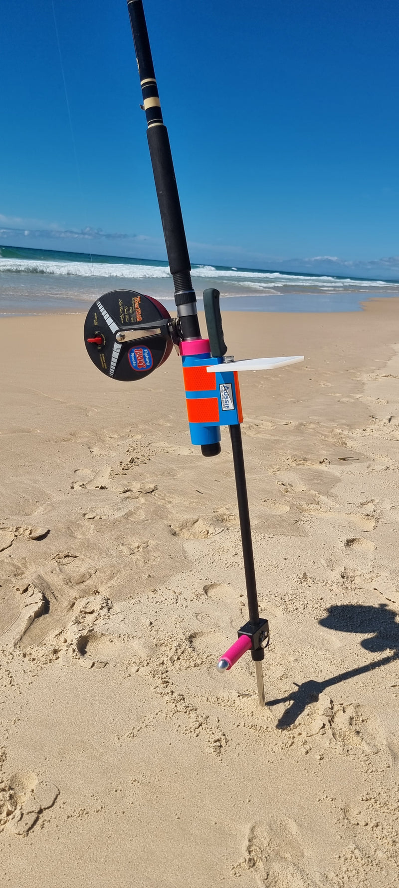 Fishing rod and reel in fibreglass fishing rod holder with bait board on a sandy beach with ocean in the background